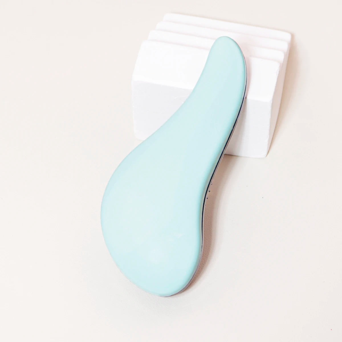 Brosse à cheveux de couleur verte posée sur un plan crème et appuyée sur un bloc décoratif blanc