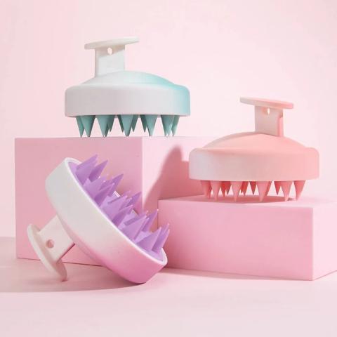 Les trois coloris de brosse cheveux massage posés sur un plan rose et des blocs décoratifs roses 