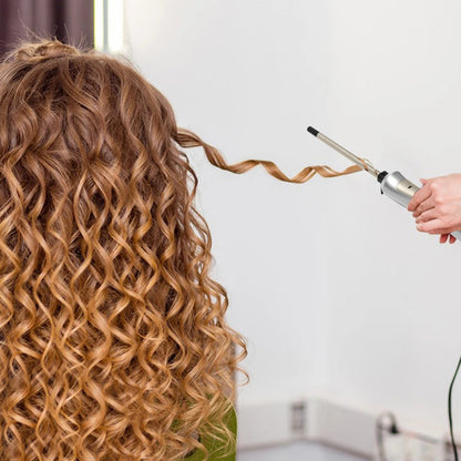 professionnelle de la coiffure qui coiffe une jeune femme aux longs cheveux bouclés châtains blonds avec le fer bouclant 
