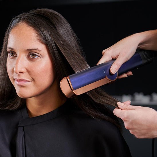 professionnelle de la coiffure qui coiffe une jeune femme aux longs cheveux bruns avec le fer bouclant de couleur bleue 