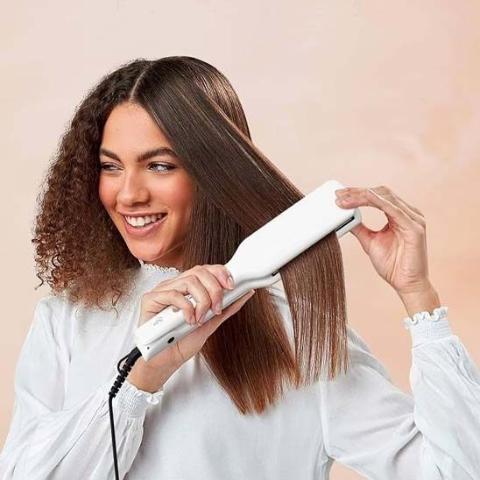 Jeune femme souriante aux cheveux mi long bouclés châtains bruns qui se coiffe avec le lisseur
