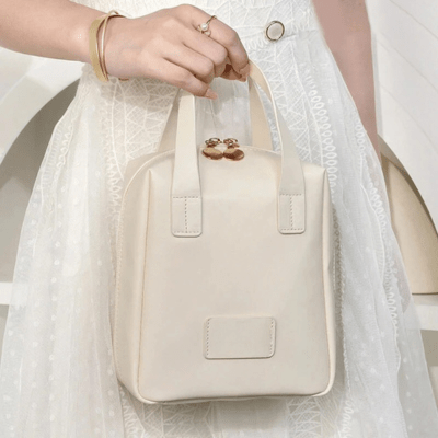 Une femme qui tient sa trousse de toilette sac à main couleur crème devant elle afin d'en apprécier les caractéristiques